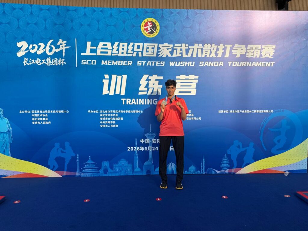 Young man in red polo stands on a blue training stage with medals, posing for a photo in front of a Chinese martial arts tournament banner, 2016 Wushu event poster.