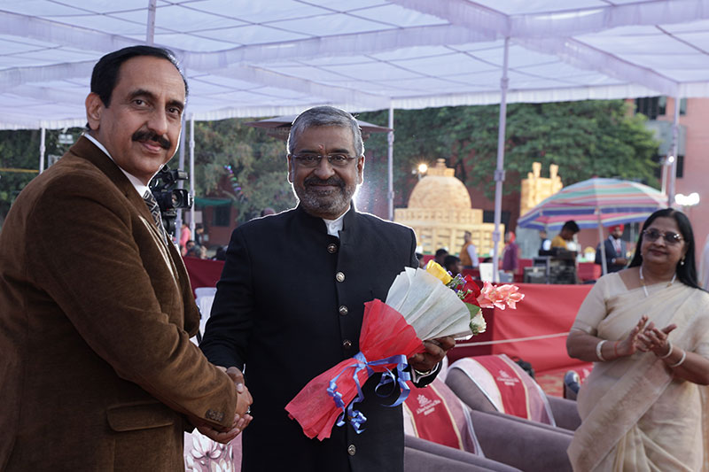 Two men shake hands at an outdoor ceremony; one holds a bouquet wrapped in red and white paper.