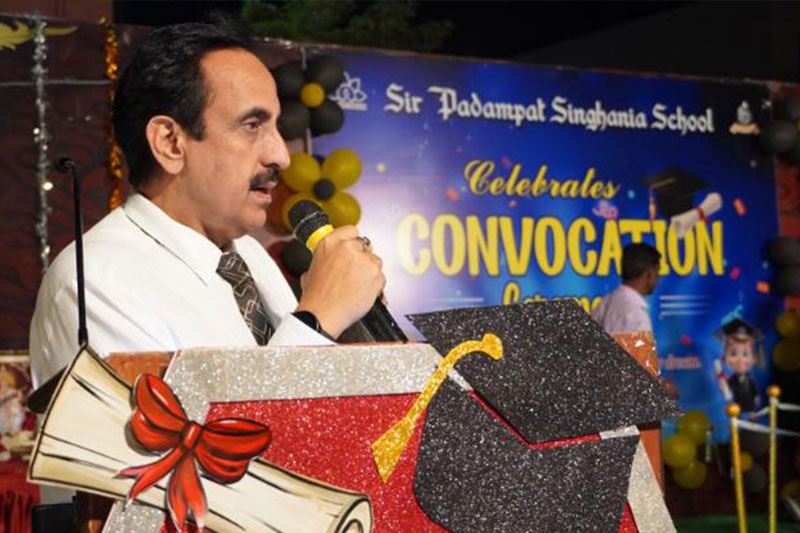 Man in a white shirt and tie speaks into a microphone at a school convocation ceremony; backdrop reads 'Sir Padampat Singhania School Celebrates Convocation'.