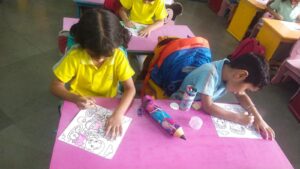 Two children color cartoon worksheets at pink tables in a classroom; one in a yellow shirt, the other in blue, with backpacks nearby.