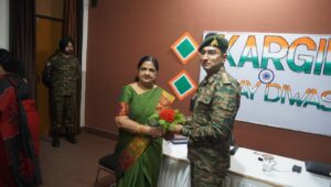 Woman in a green saree hands a bouquet to a soldier in camouflage at a Kargil Vijay Diwas event indoors, banner in background.