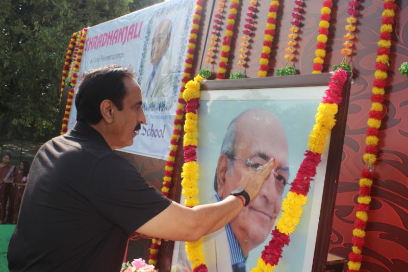 Man applying a tilak to a framed portrait garlanded with marigolds at an outdoor tribute ceremony.