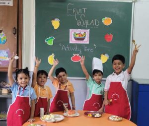 Group of smiling children in colorful shirts and red aprons raise their hands in a classroom, with a fruit salad activity setup on an orange table and a chalkboard that says 'Fruit Salad Activity Time' surrounded by fruit cutouts.