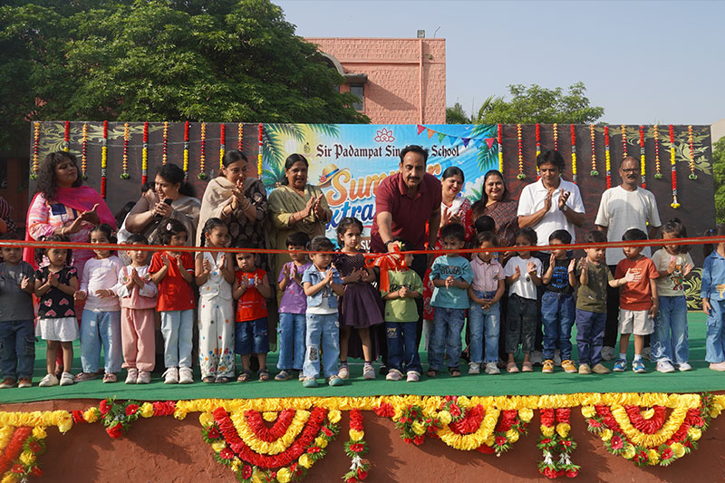 School event with a line of children and adults cutting a red ribbon on a decorated stage, festive banners and flowers in the background.